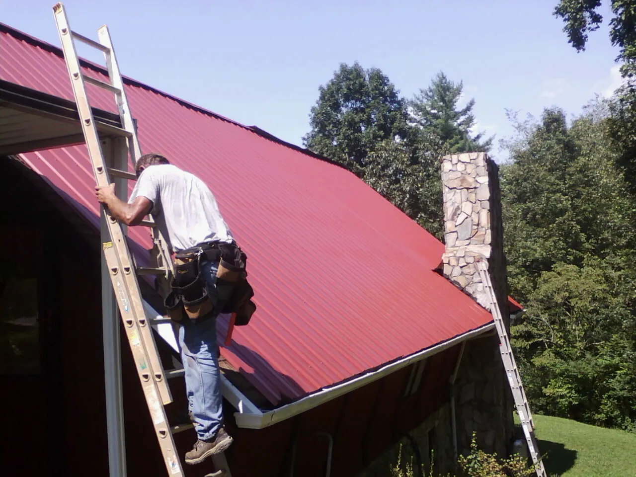 Roofer on a ladder beside a red metal roof and stone chimney — Soffit & Fascia Repair by Roofing Brothers in New Middletown